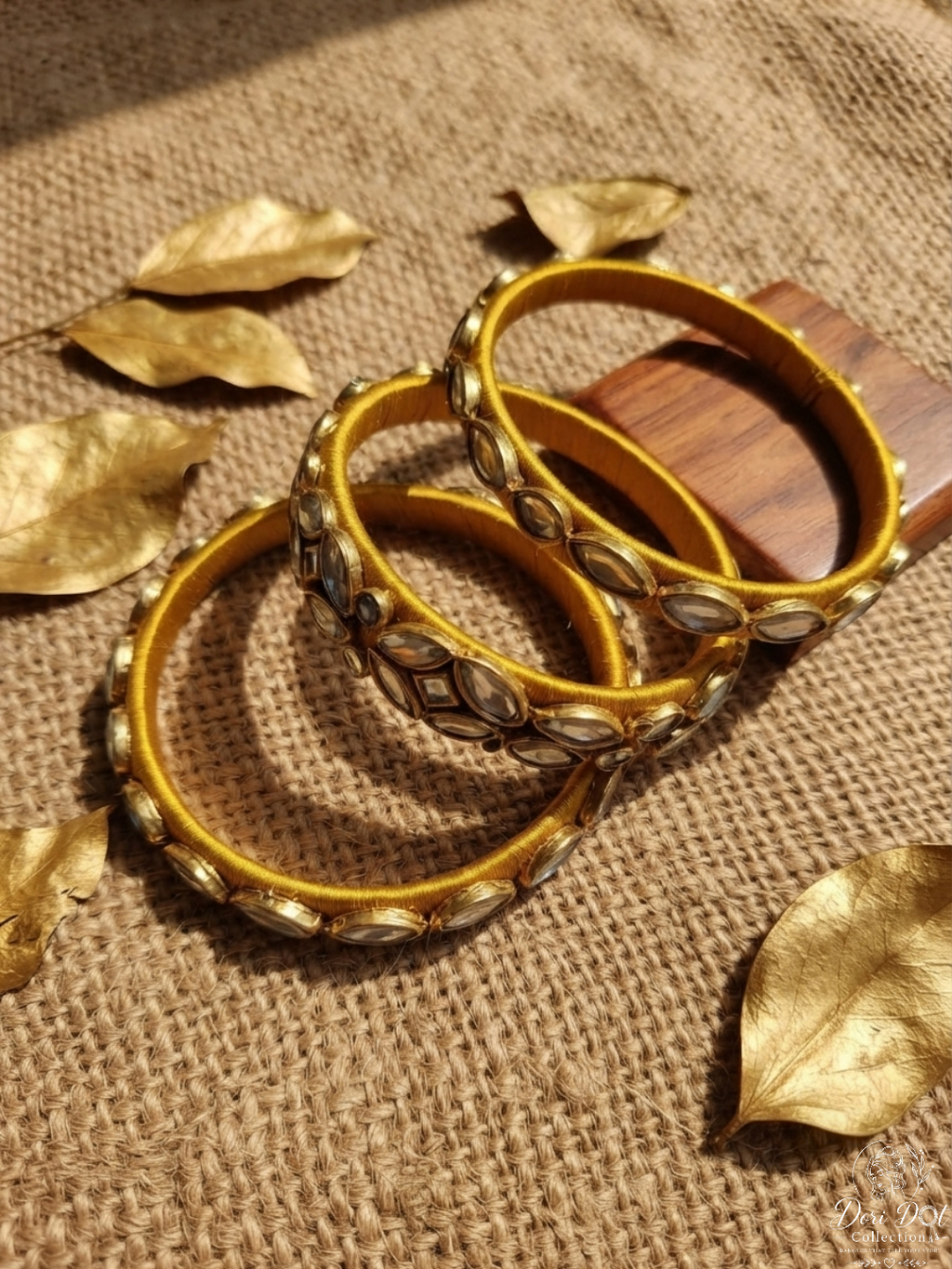 Three gold bangles adorned with silver-toned oval stones, arranged on a textured beige burlap surface with scattered gold-toned decorative leaves.