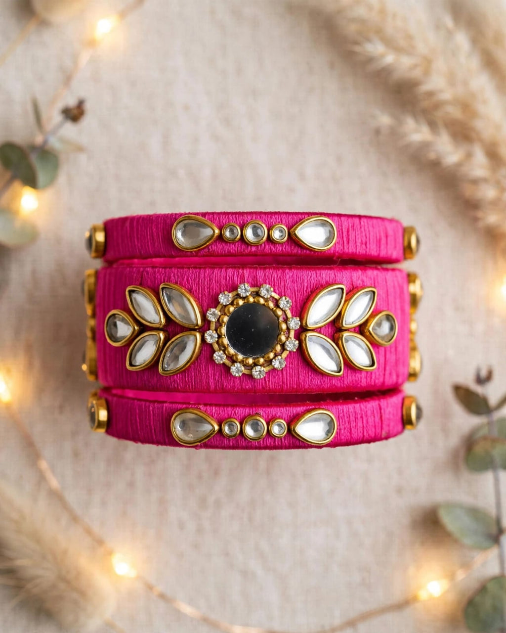 A bright pink fabric cuff bracelet with gold-toned metal accents, white mirror-like stones, and a central black stone, styled on a beige textured surface with warm fairy lights and dried grass.