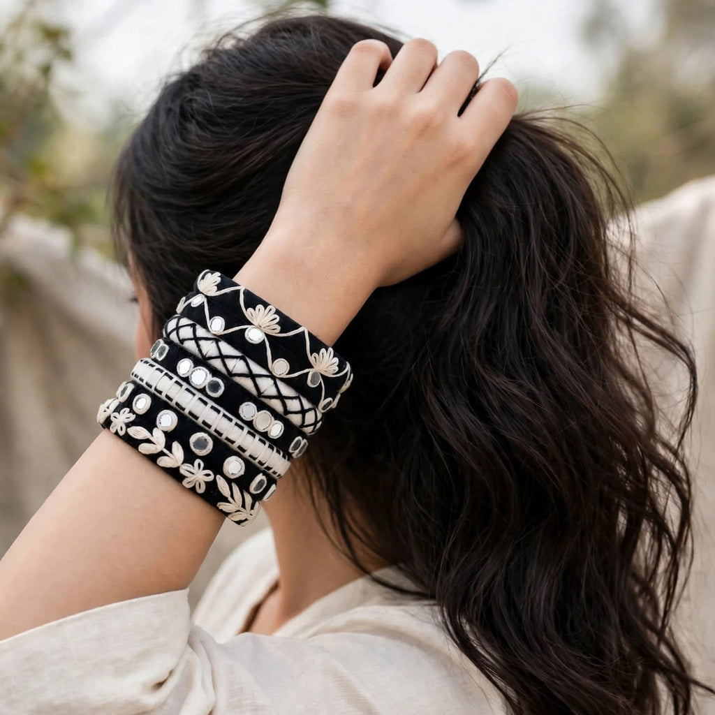 A woman with long dark hair seen from the back, wearing a wide black cuff bracelet with white floral embroidery and bead details on her wrist, set against a soft-focus outdoor background.