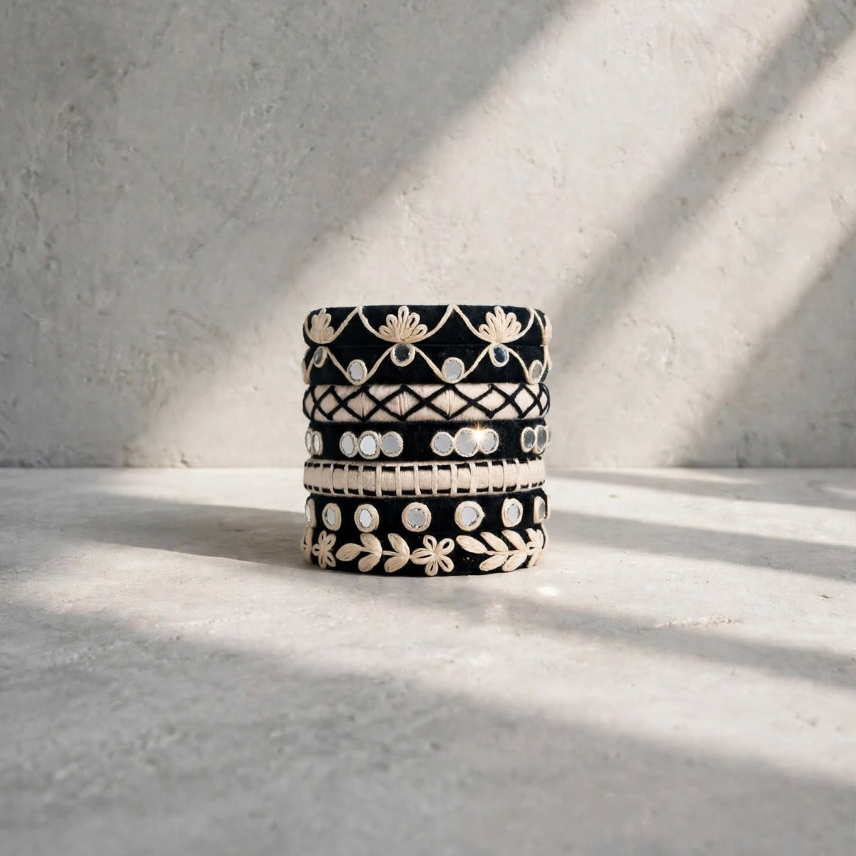 A stack of four black and white patterned bangles with intricate floral and geometric designs, styled on a textured light gray surface under soft diagonal sunlight.