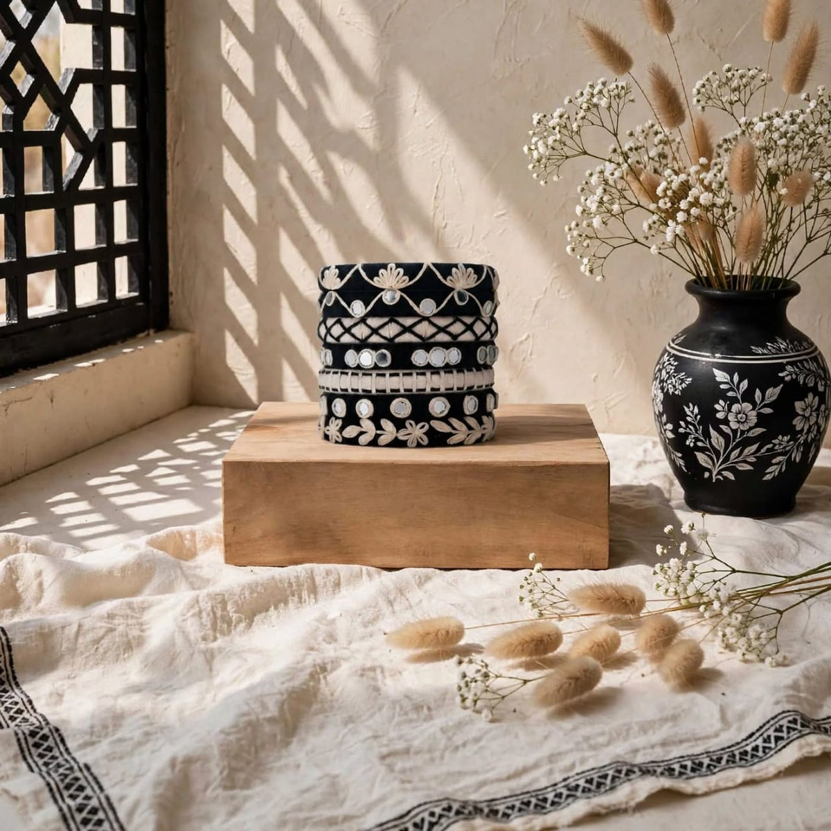 A stack of three black and white patterned ceramic boxes sits on a wooden block on a textured cream cloth, styled with a matching black floral vase of dried grasses and baby's breath in a sunlit room.